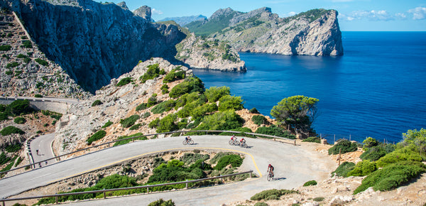 Three cyclists cycling along a road in Mallorca, Spain, that leads to Formentor lighthouse with the Mediterranean sea in the background 